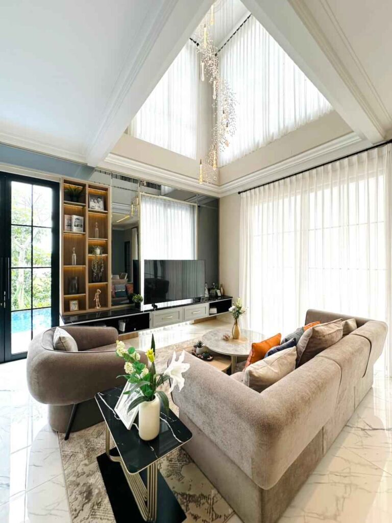 bright living room with a beige sectional sofa marble floor and large windows with sheer white curtains tv and built in shelving unit   citragran cibubur | perumahan baru lokasi strategis cibubur